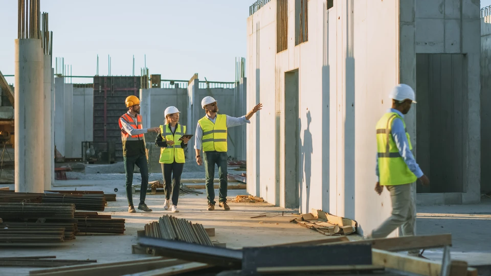 Chantier de construction intérieur avec trois professionnels du bâtiment en équipements de protection individuelle (casques jaune, bleu, blanc, gilets haute visibilité) analysant des plans architecturaux sur table de travail temporaire